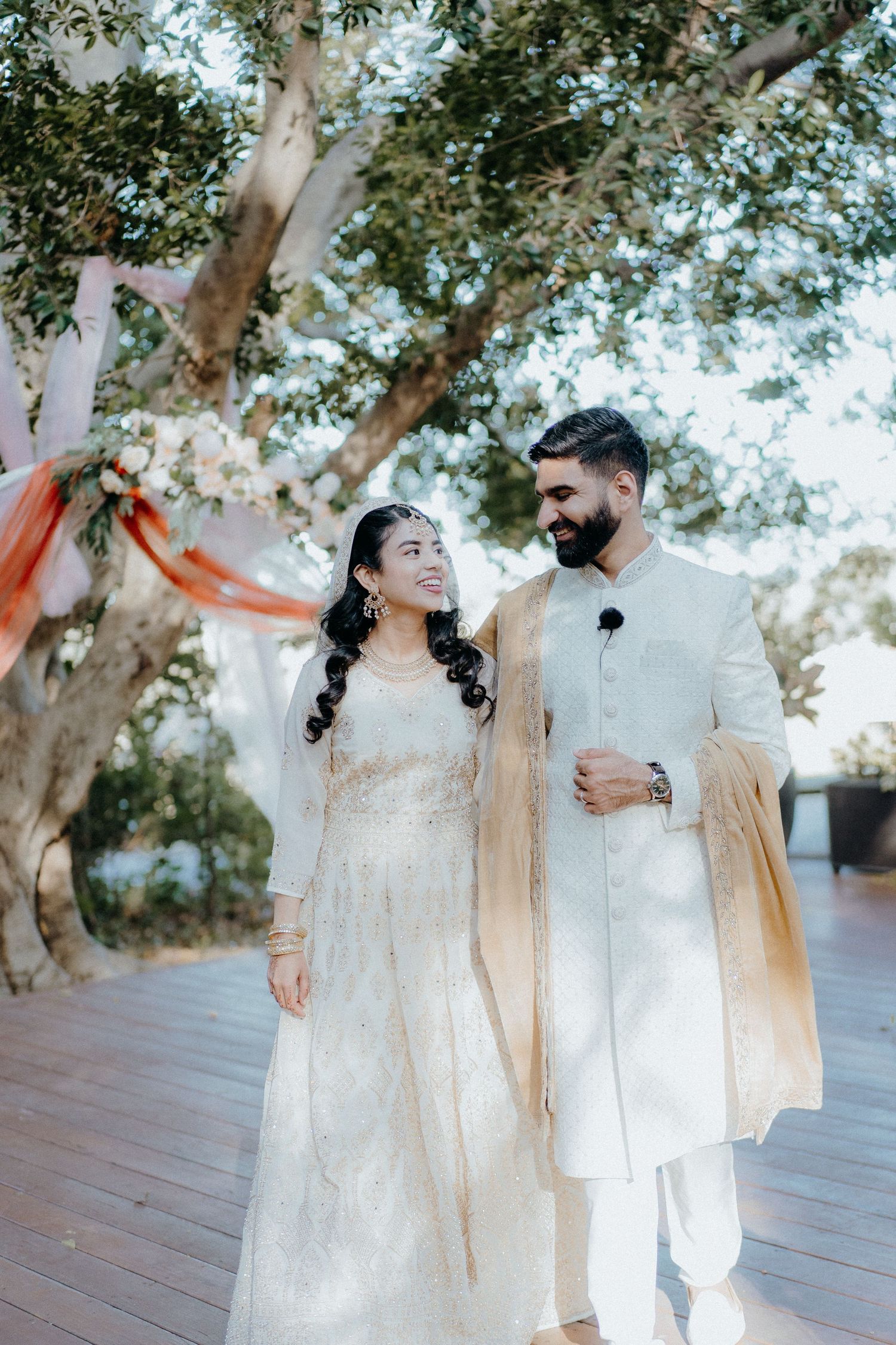 Elegant couple in white traditional wedding attire walking under decorated tree canopy.