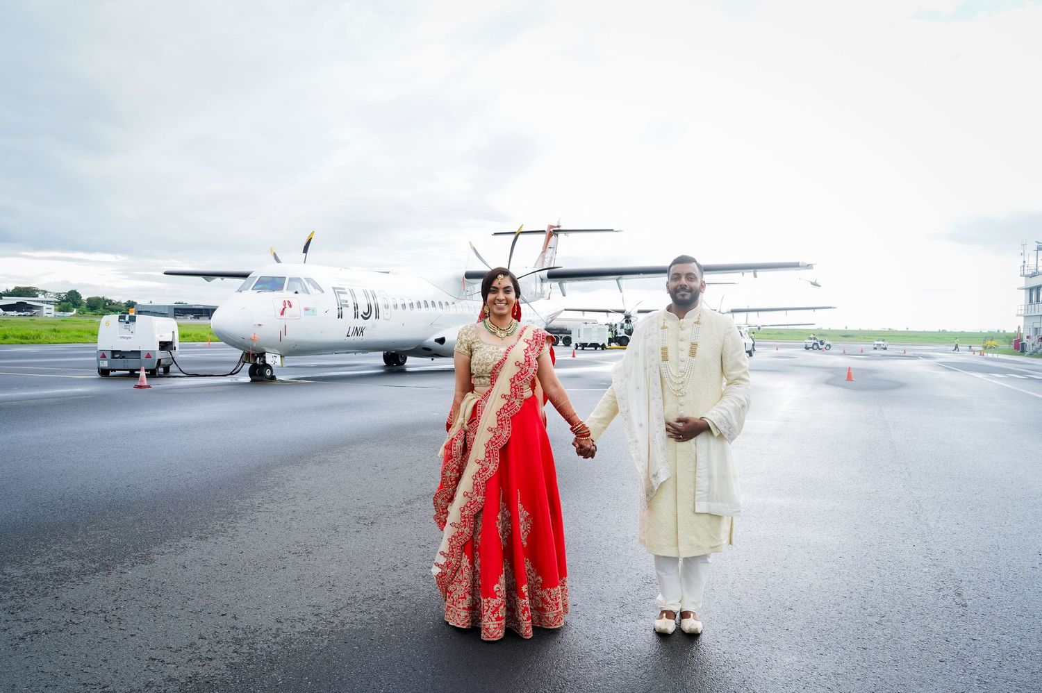 Newlyweds in traditional Indian wedding attire pose near small aircraft on runway.