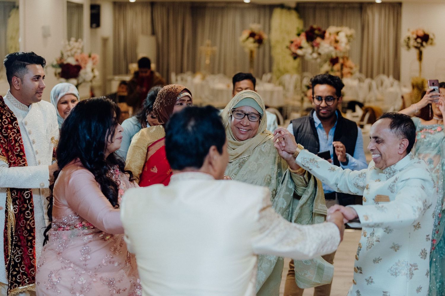 Wedding guests circle dancing together in formal ethnic attire at an indoor celebration.