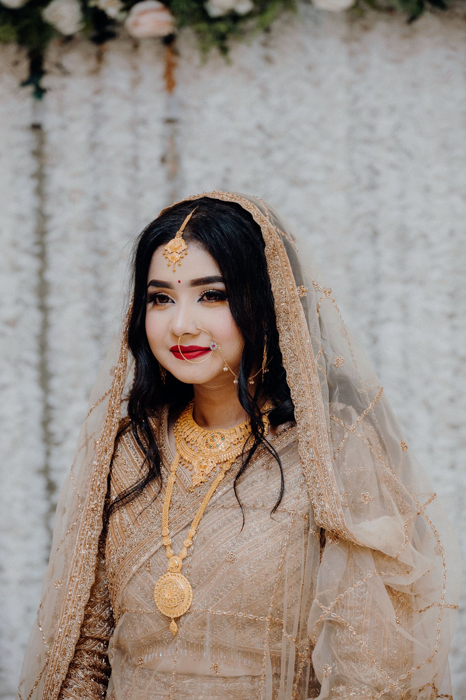 Bride wearing gold jewelry and beige veil poses elegantly against floral backdrop.