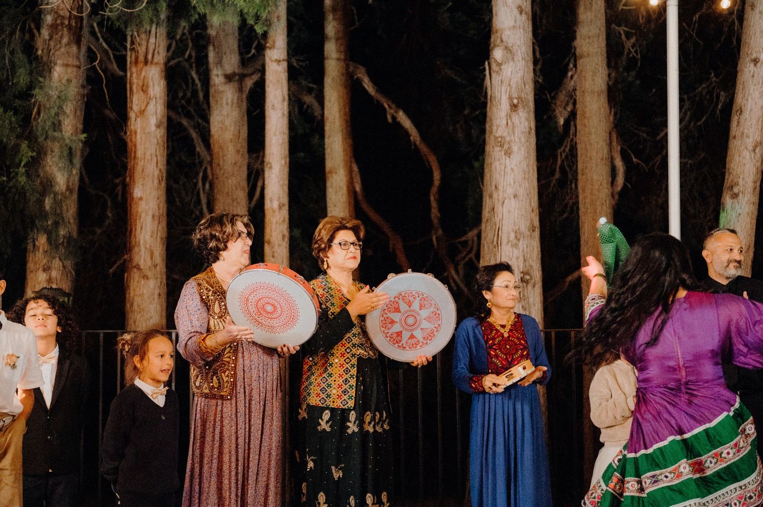 People in traditional folk costumes holding decorative parasols pose in front of a forest backdrop.