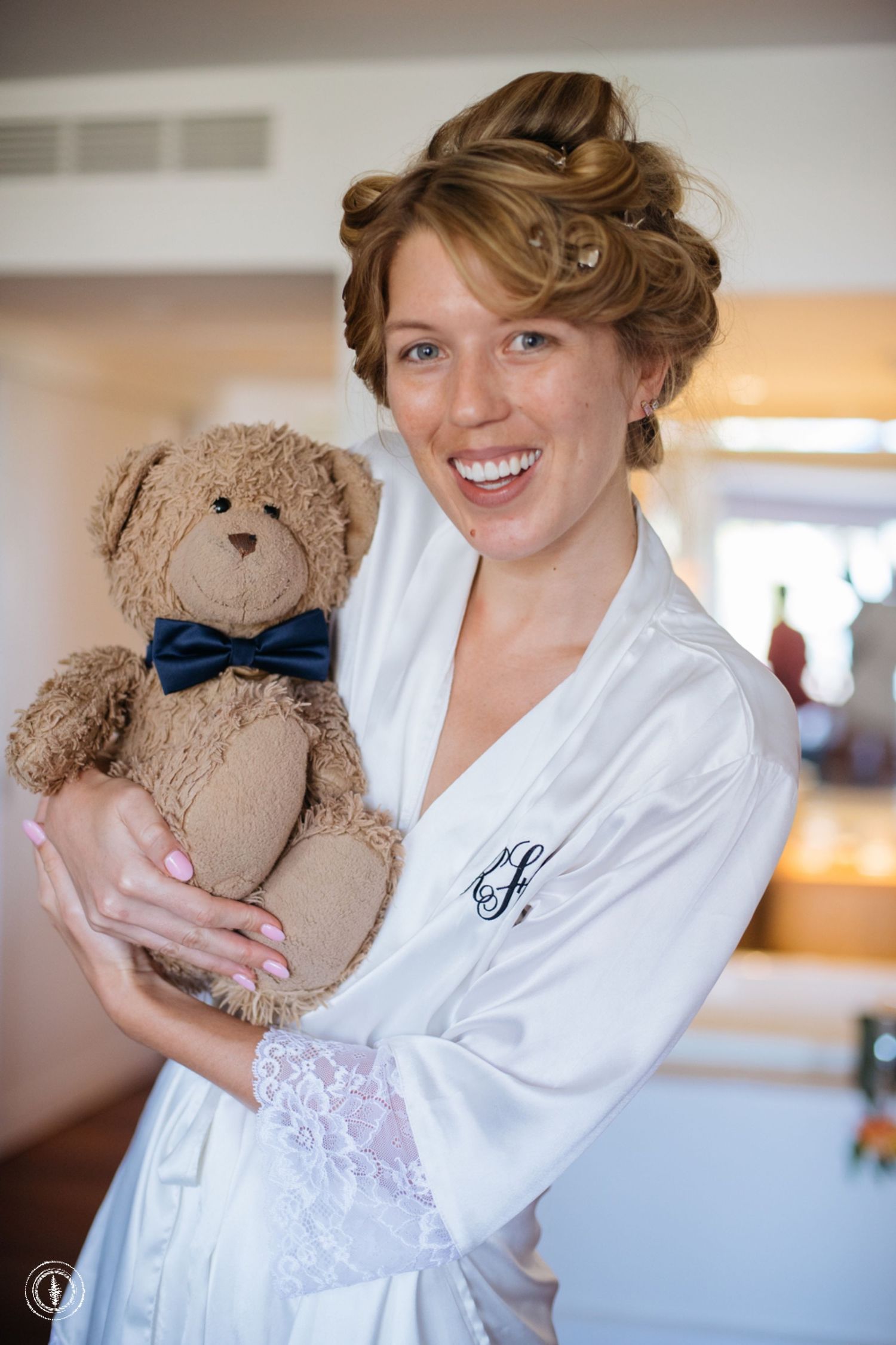 A person in a white robe holding a teddy bear with a blue bowtie during wedding preparations.