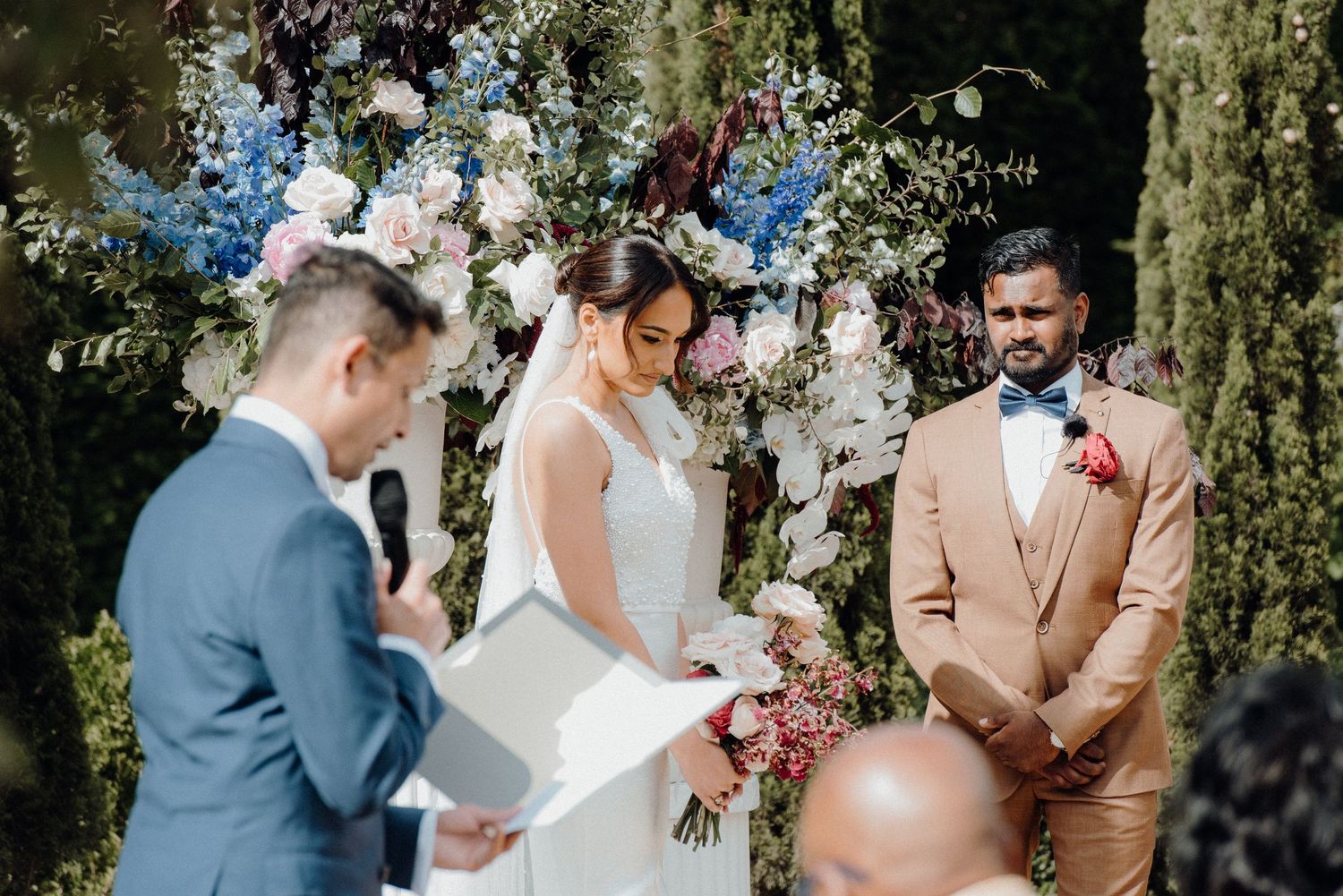 Outdoor wedding ceremony features an elegant floral arch with blue and white blooms against cypress trees.