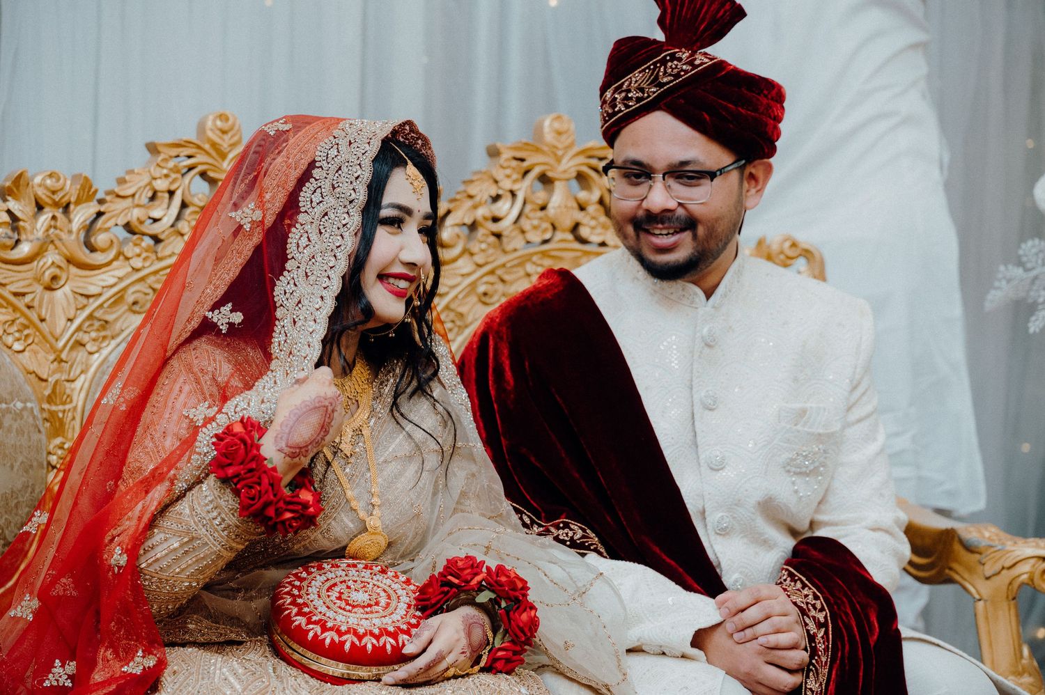 A wedding couple sits together on an ornate golden throne wearing elegant South Asian ceremonial dress.