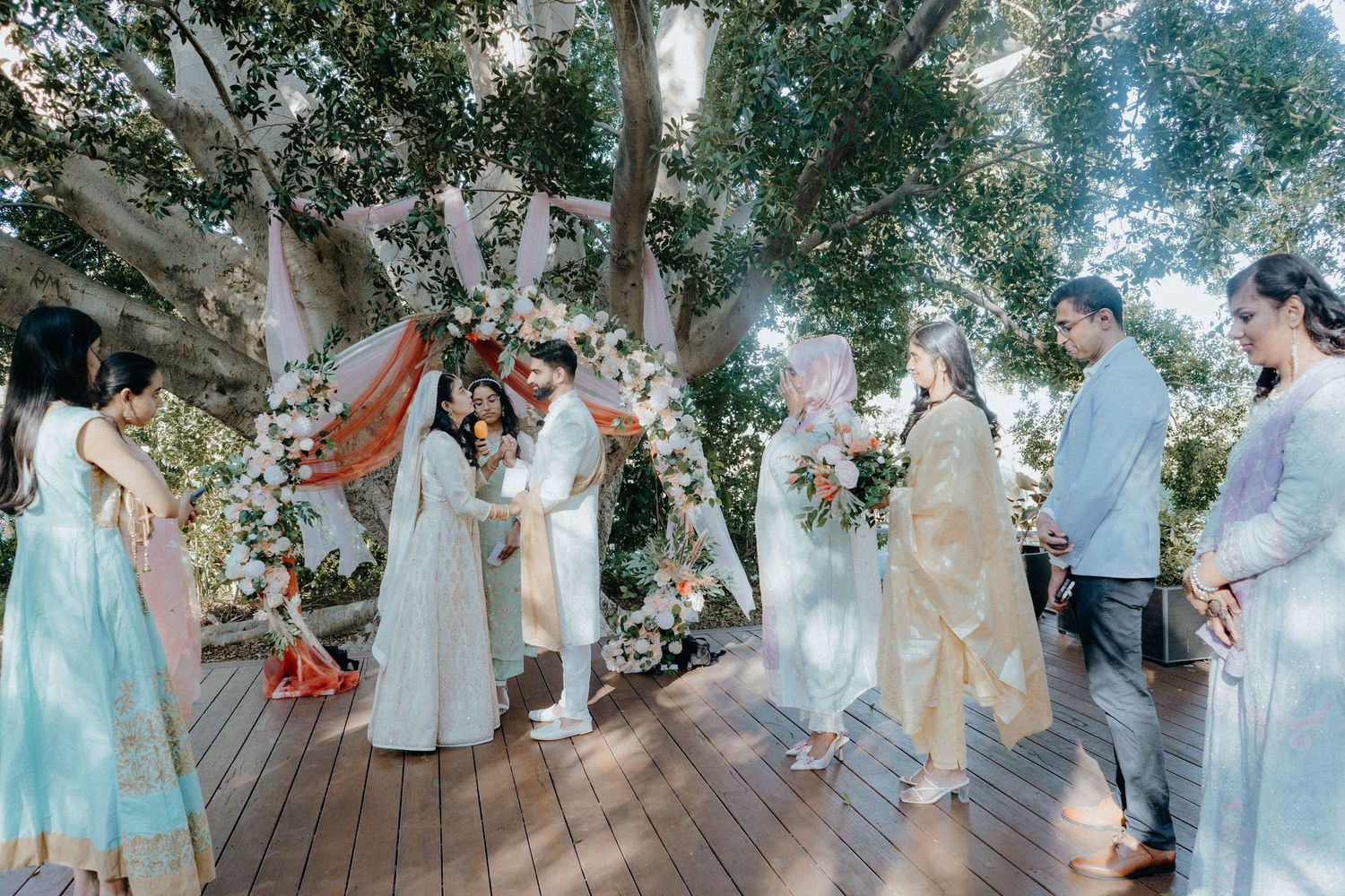 A beautiful outdoor wedding ceremony setup with floral decorations and white draping under a canopy of trees.