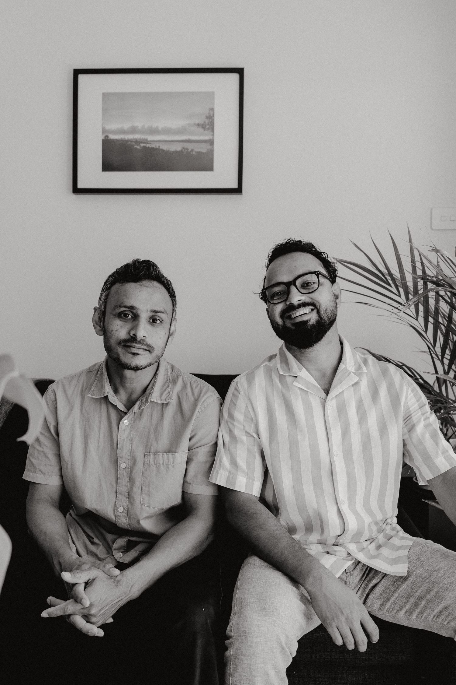 Black and white portrait of two smiling men sitting on a couch beneath a framed landscape photo.