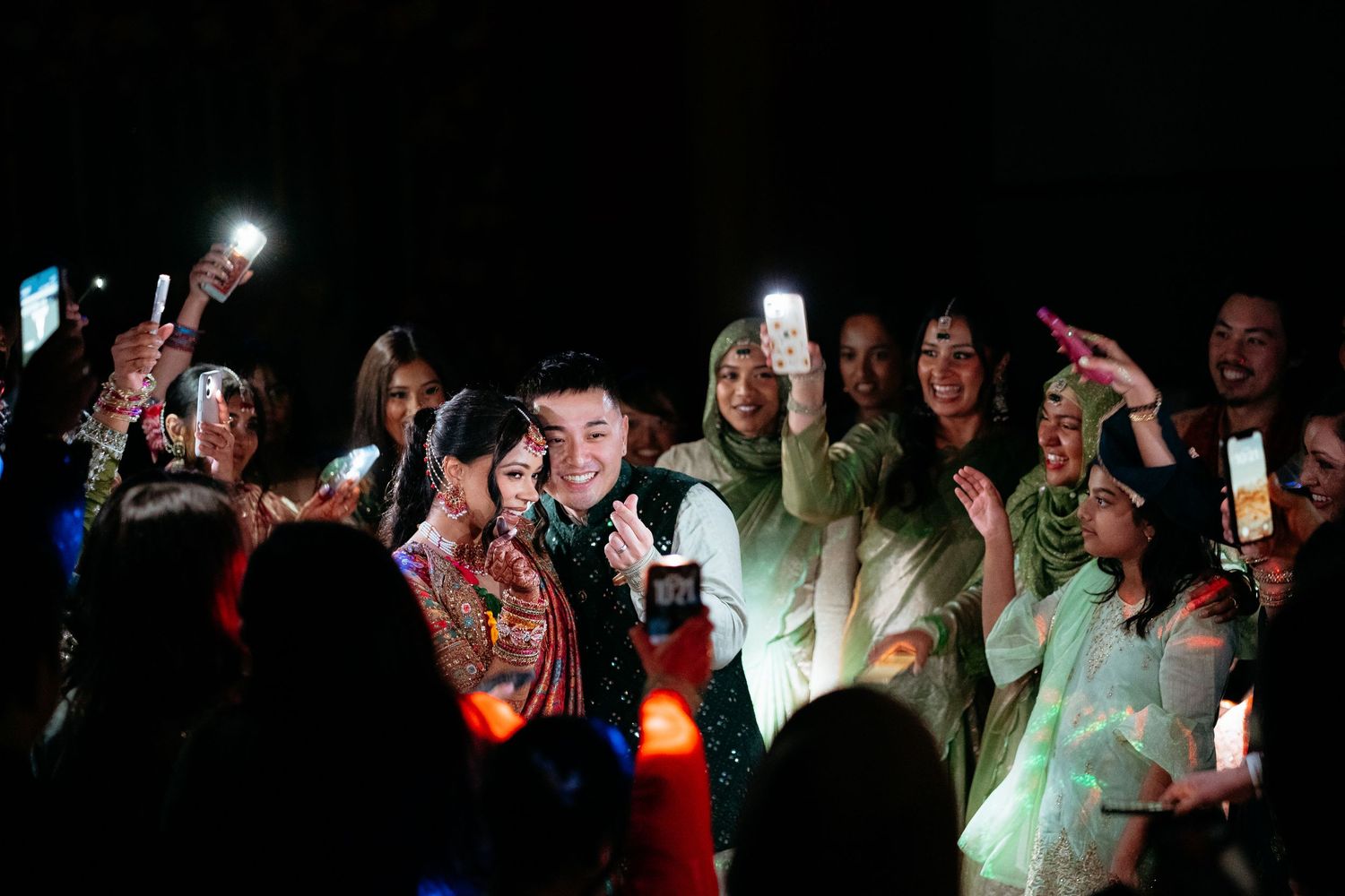 Wedding couple surrounded by celebrating guests holding phone flashlights at a vibrant nighttime South Asian wedding reception.