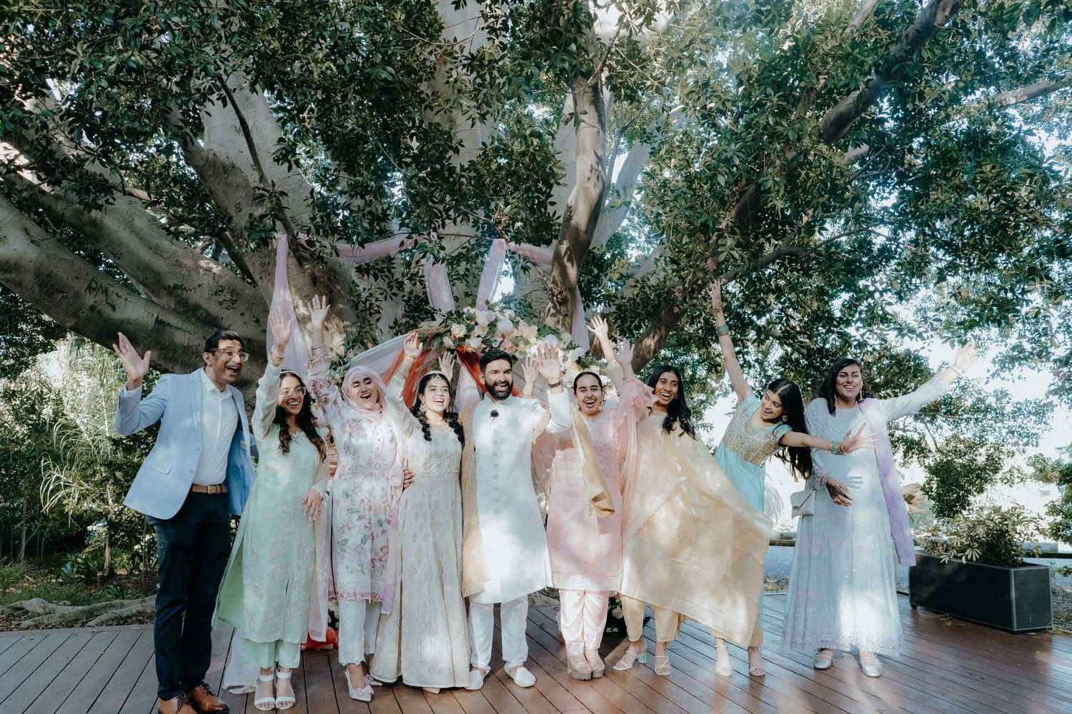 Wedding party in coordinated pastels and whites celebrating outdoors under tree canopy.
