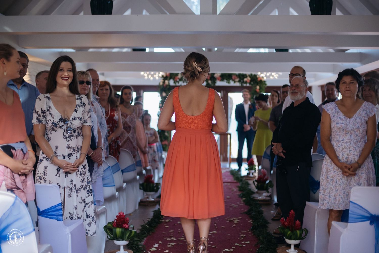 Guest in coral dress walking down the aisle at an indoor wedding ceremony.
