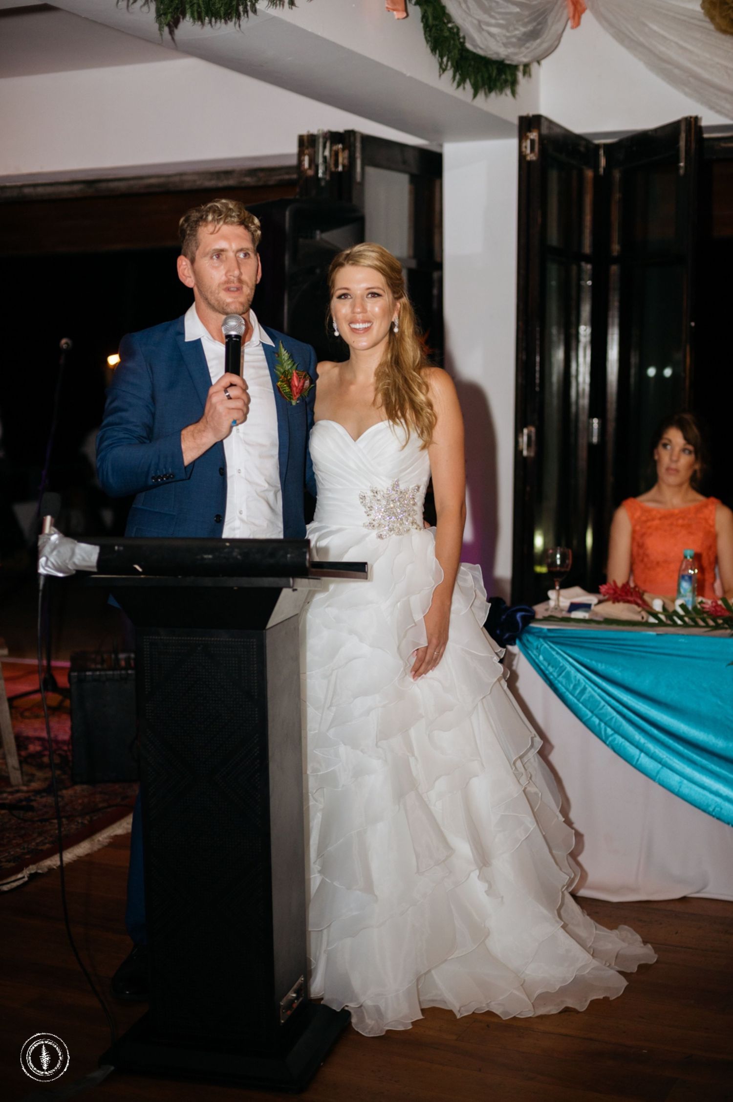 Wedding couple stands at podium delivering reception speech with elegant decor in background.