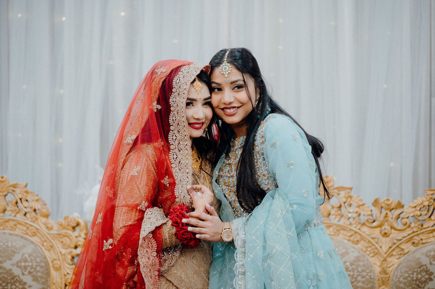 Two guests in traditional South Asian attire pose together at a wedding celebration against a light backdrop.