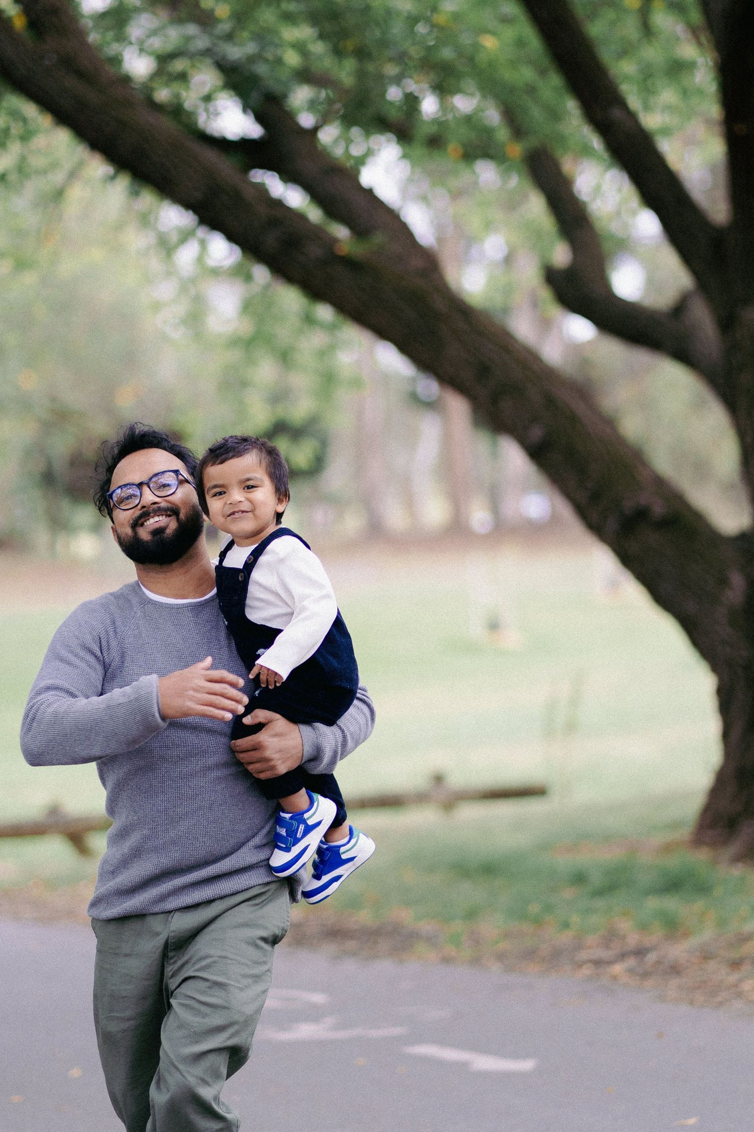 A bearded father carrying his smiling toddler while walking through a scenic green park.