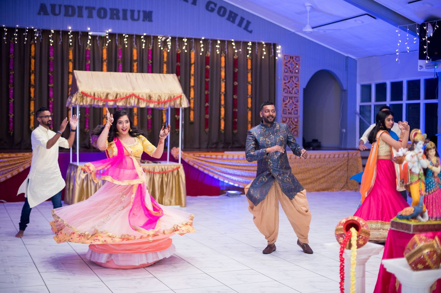 Wedding guests dancing energetically in a decorated auditorium with colorful lights and decorations.