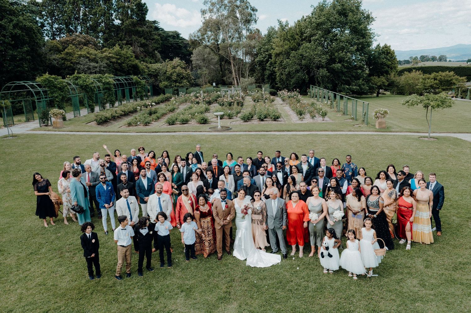 Large wedding group photo taken from above on a manicured lawn with formal gardens in the background.