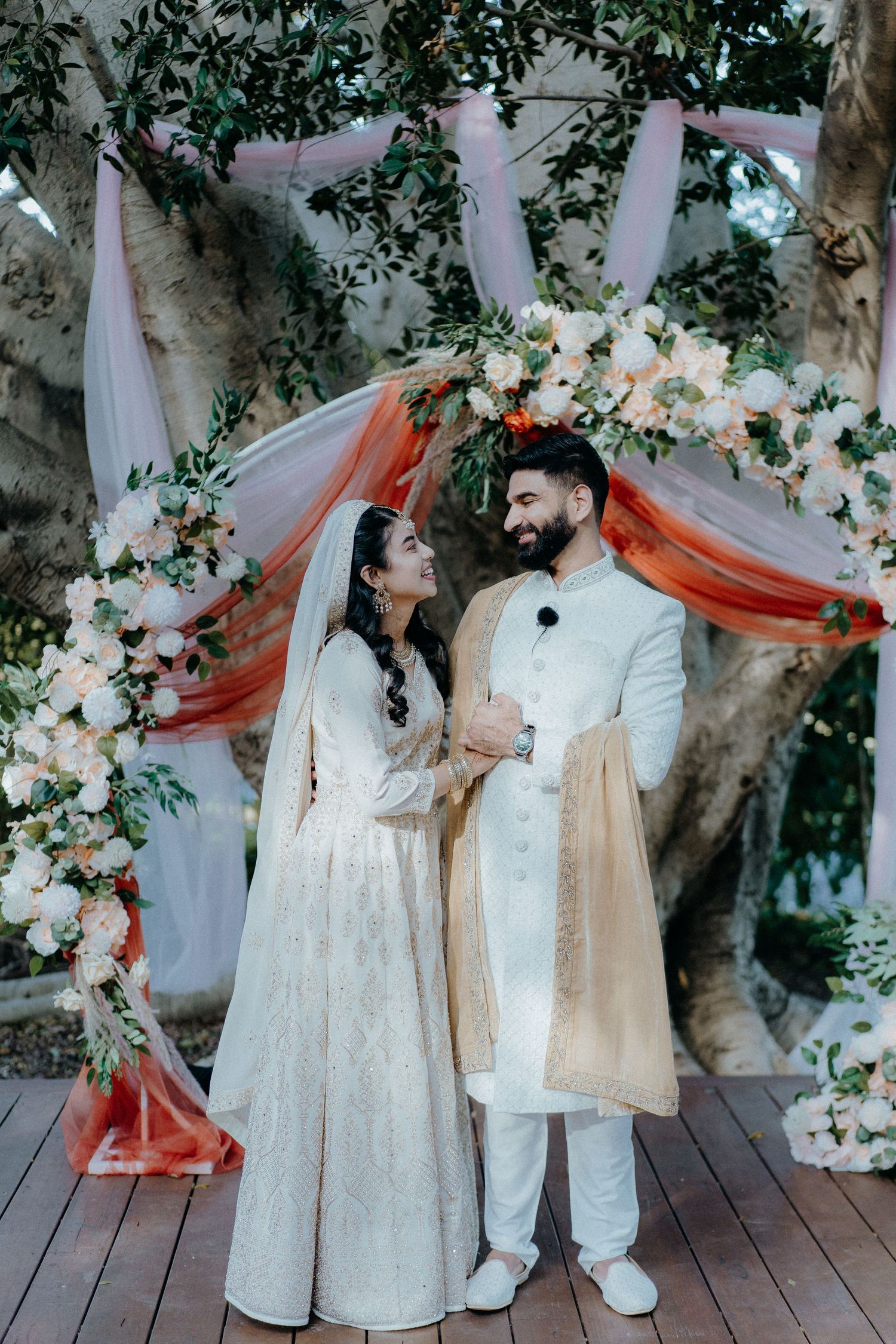 Couple in elegant white traditional wedding attire posing under a floral arch with coral drapes.
