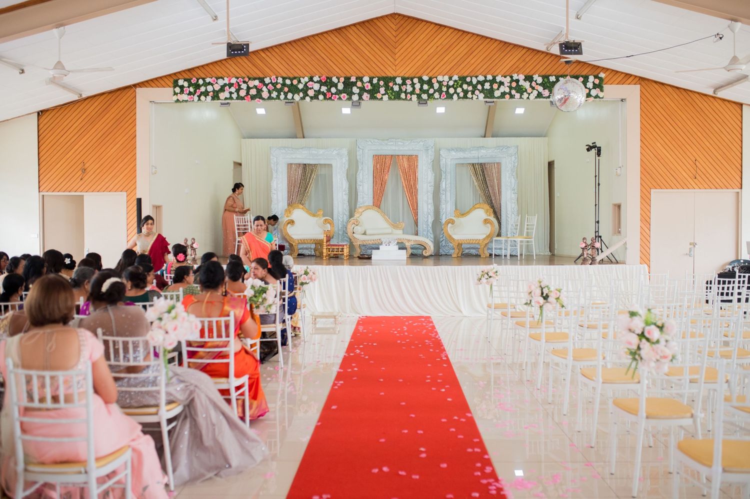 Indoor wedding ceremony setup with chairs, red carpet aisle, and floral arch decorations.