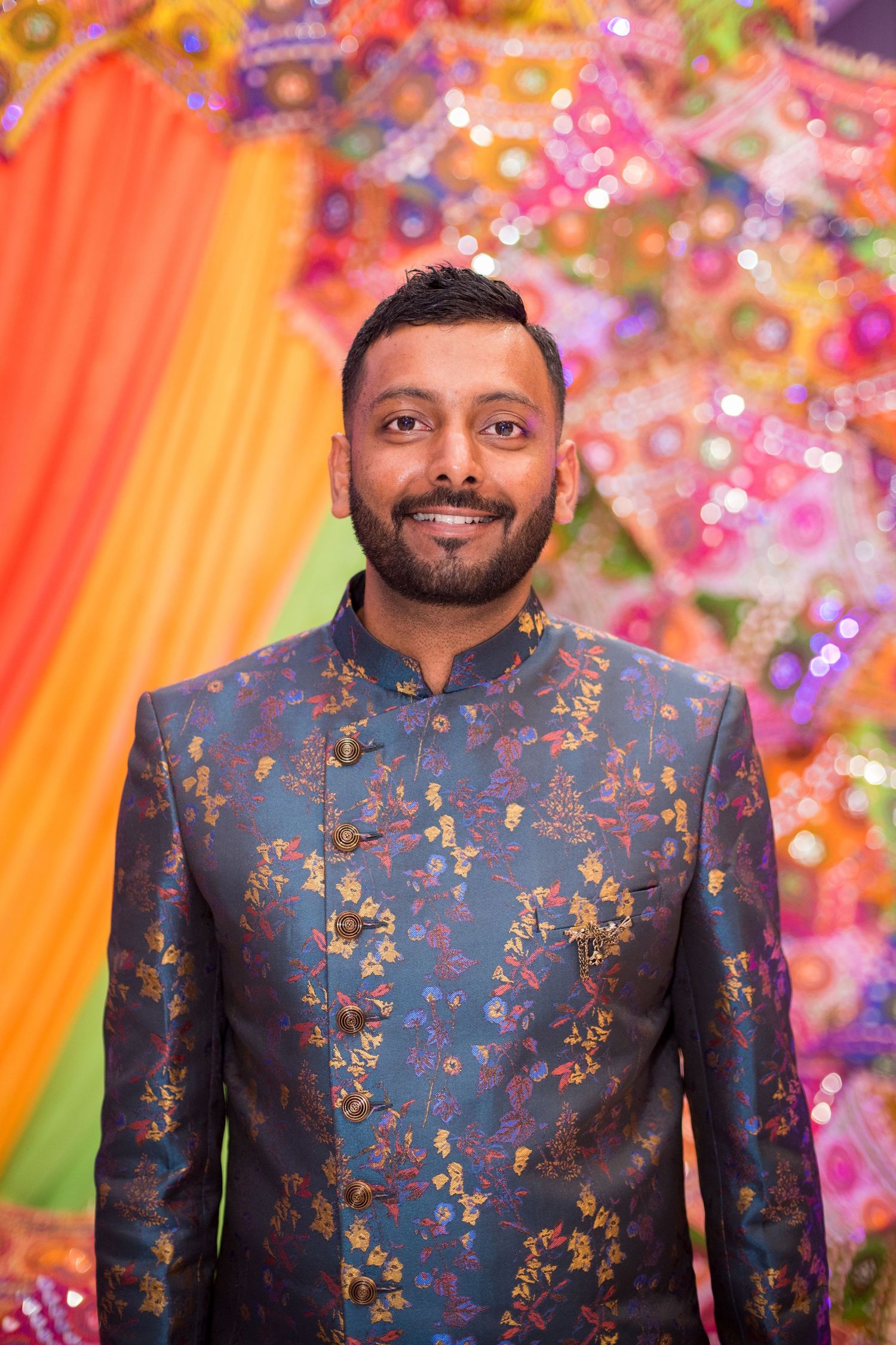 Person wearing a navy blue floral sherwani poses against a vibrant rainbow backdrop with floral decorations.