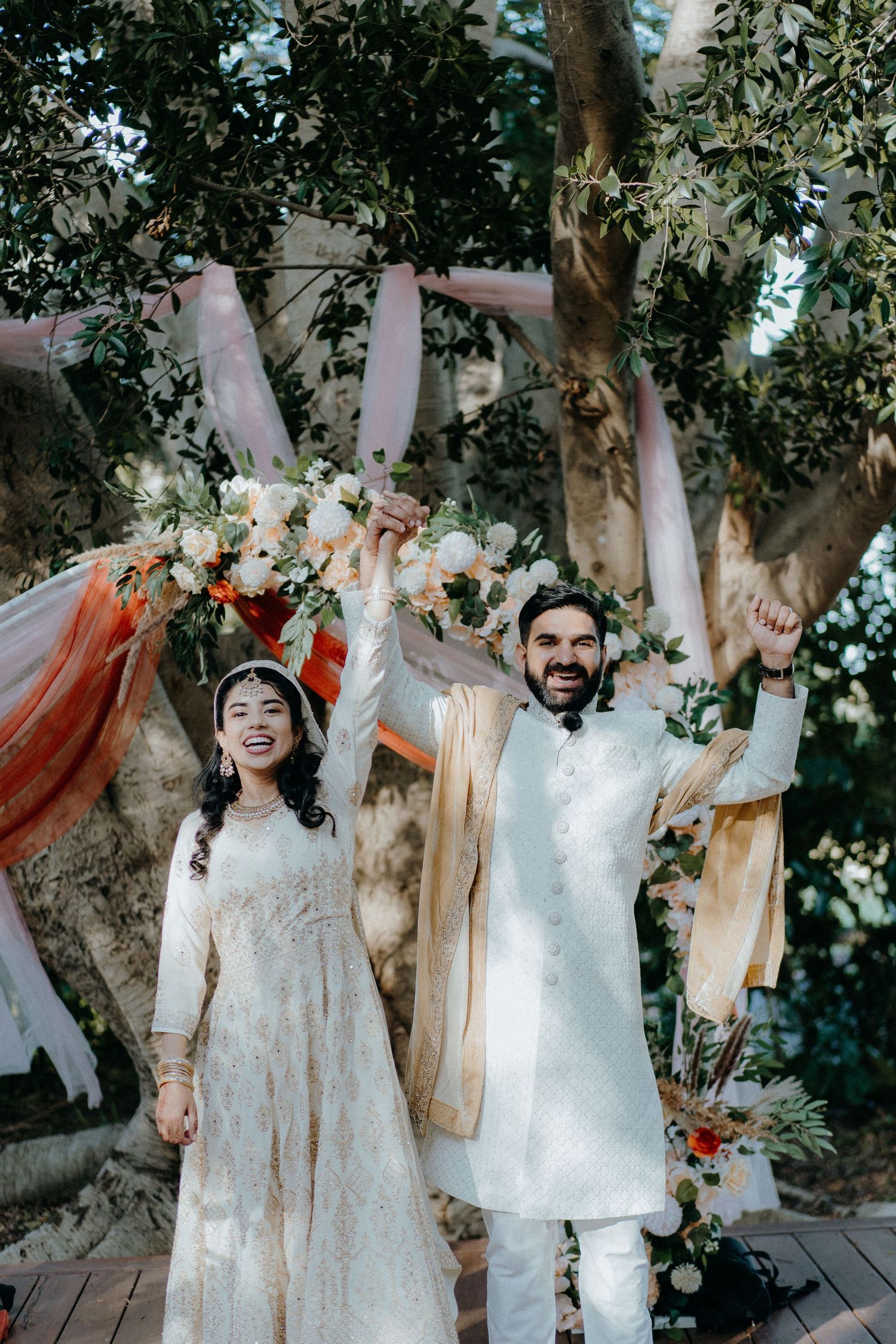 Jubilant newlyweds raise their arms in celebration under a floral-decorated garden arch.