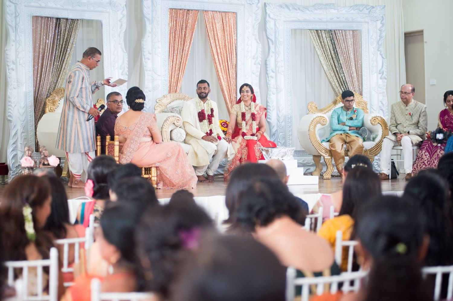 Wedding ceremony in progress with guests seated in white chairs viewing the decorated stage.