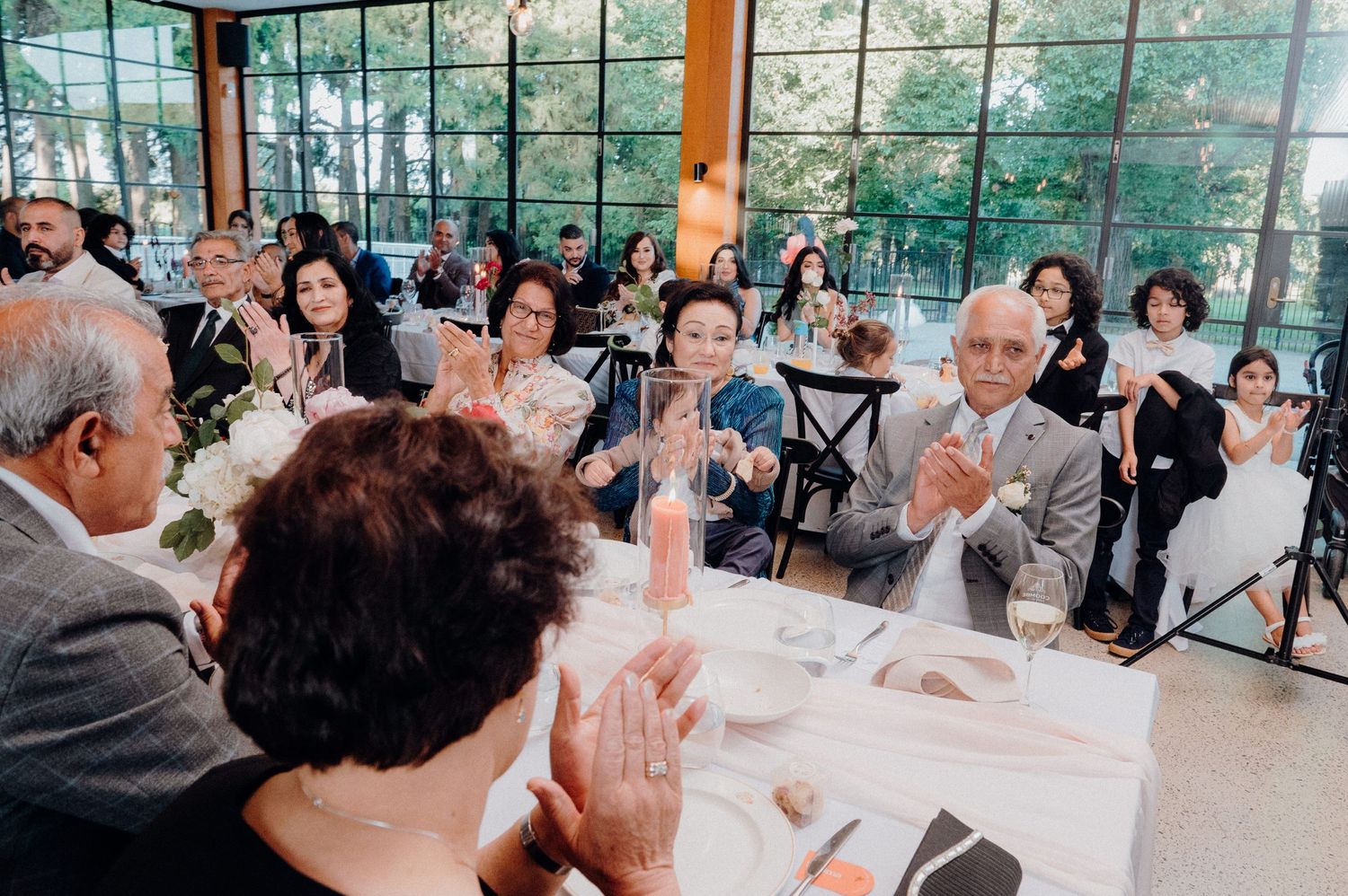 Wedding guests applauding during reception in sunlit venue with large windows.