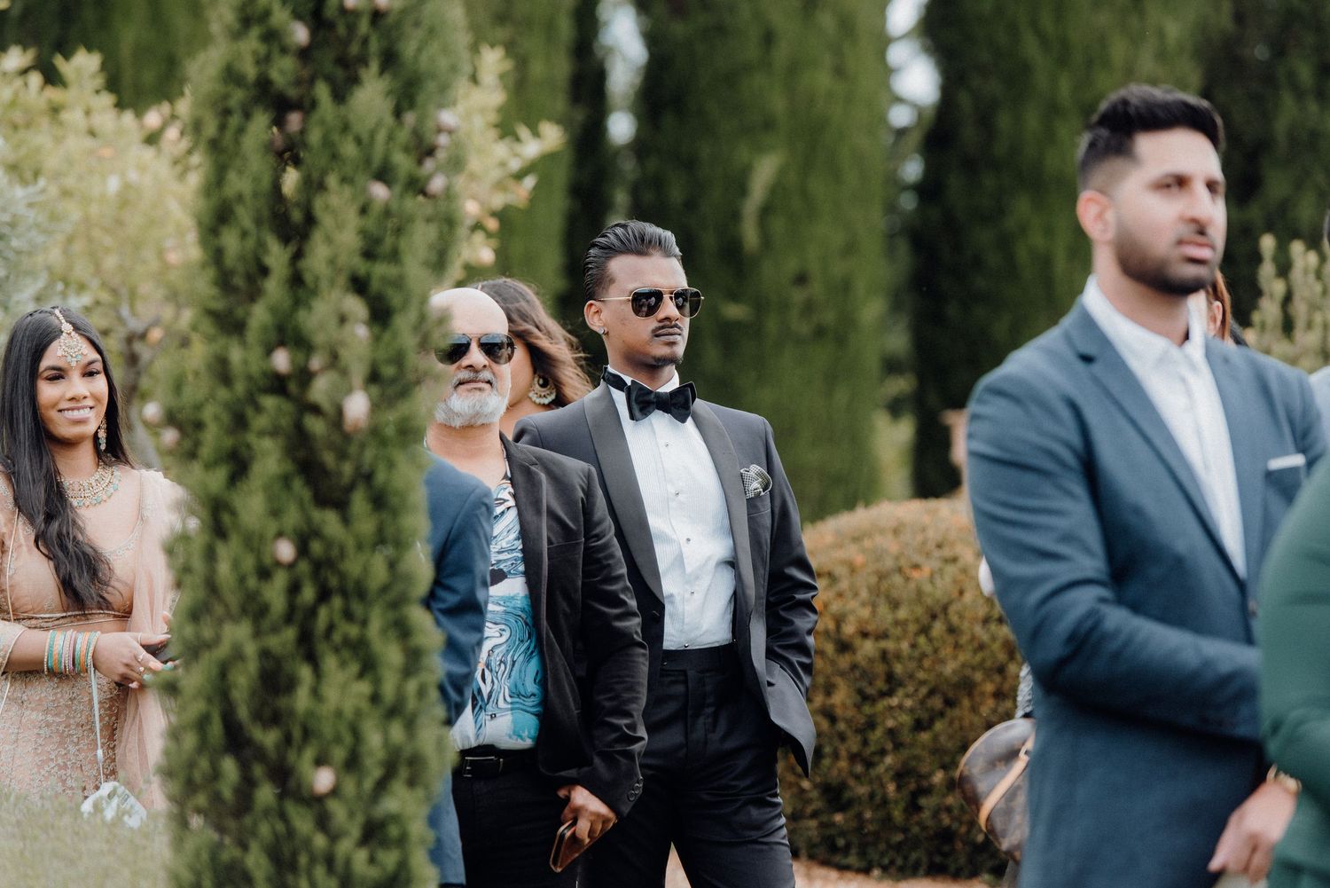 Wedding guests in formal attire standing near cypress trees during outdoor ceremony.