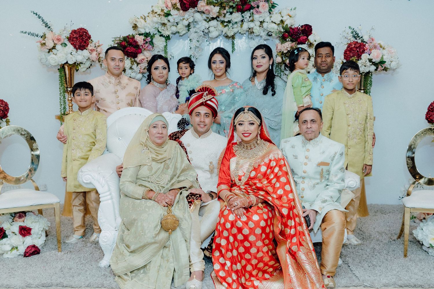 Large family group photo with wedding couple in traditional attire against floral backdrop.