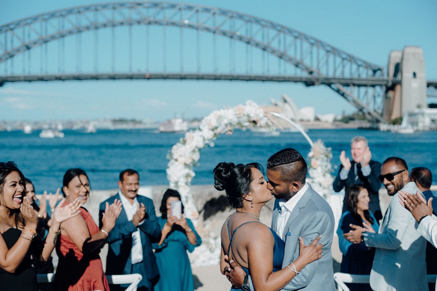 Couple shares first kiss at outdoor wedding ceremony with Sydney Harbour Bridge in background, guests applauding.