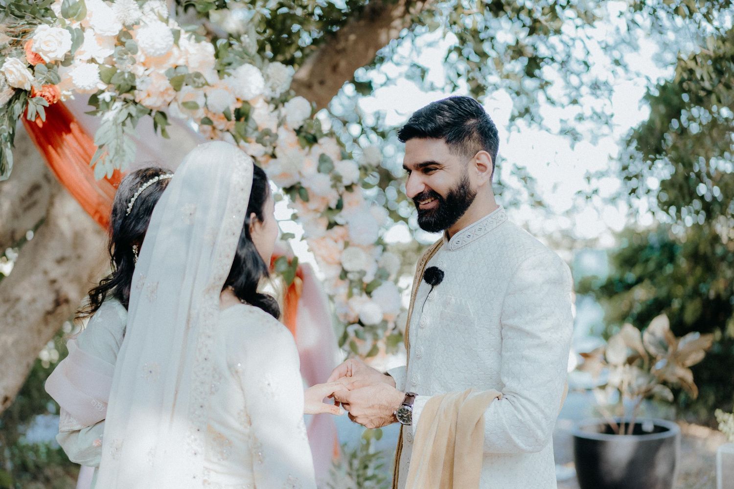 A wedding ceremony takes place under a floral arch in an outdoor garden setting.