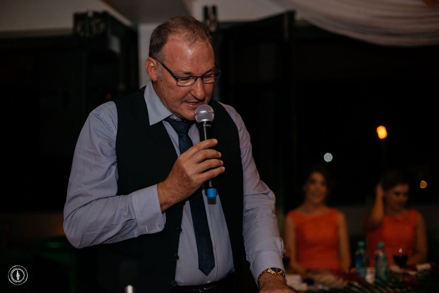 A speaker in formal attire addresses guests at an evening reception while holding a microphone.