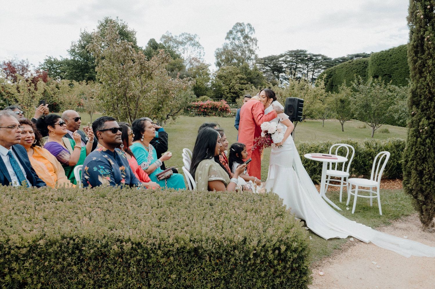 Wedding ceremony guests watching proceedings in garden setting with manicured hedges.