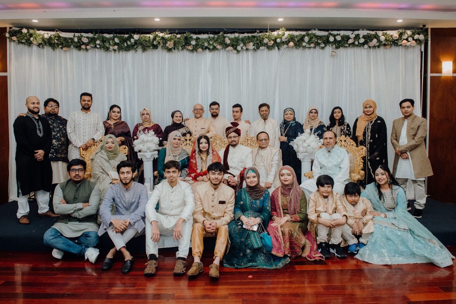 Large group photo of wedding guests in traditional South Asian attire at reception venue.