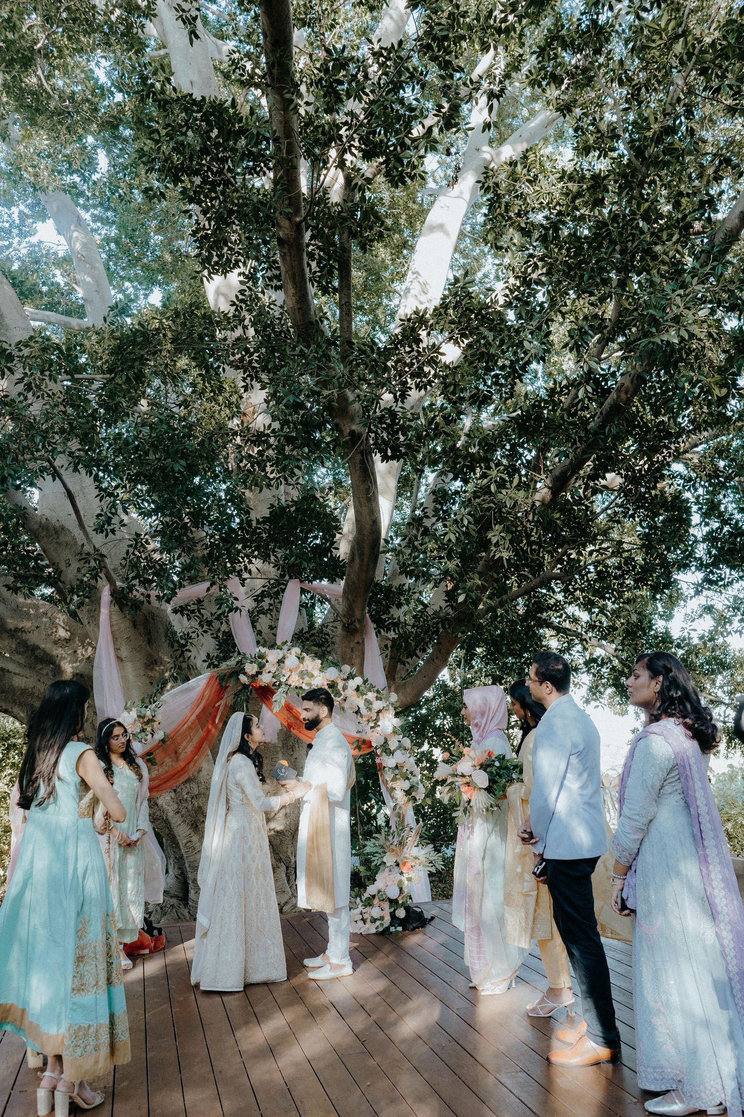 Wedding guests and couple gather on wooden deck under trees decorated with white and orange draping.