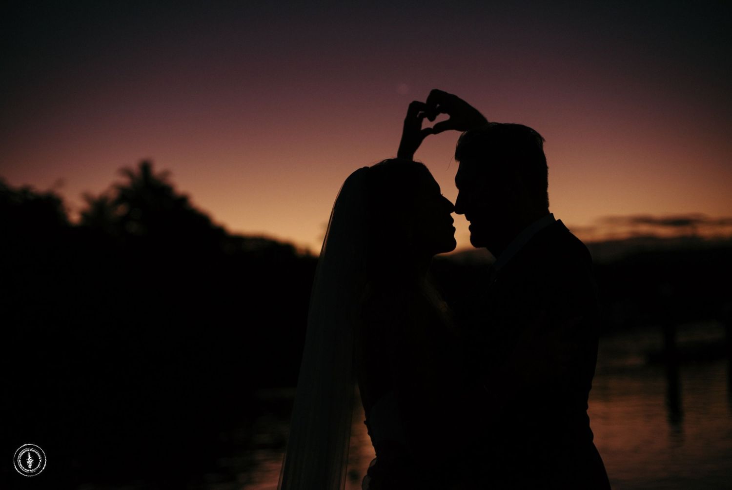 Silhouetted couple forms heart shape with hands against sunset sky.