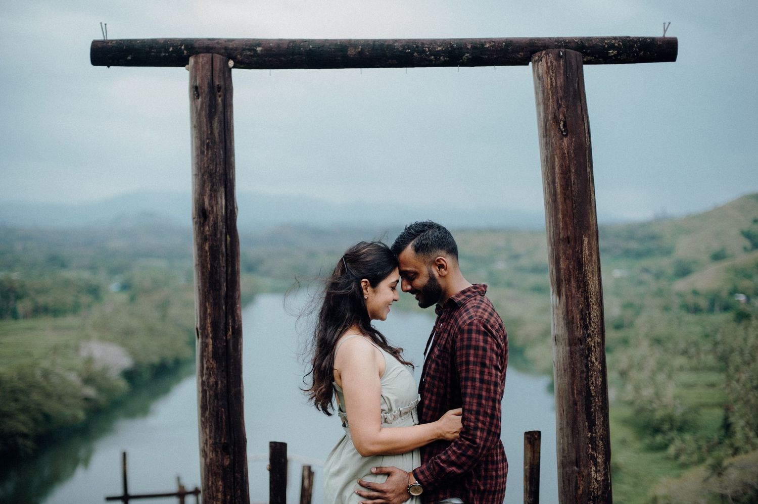A romantic embrace under a wooden arch with a winding river and mountain landscape in the background.