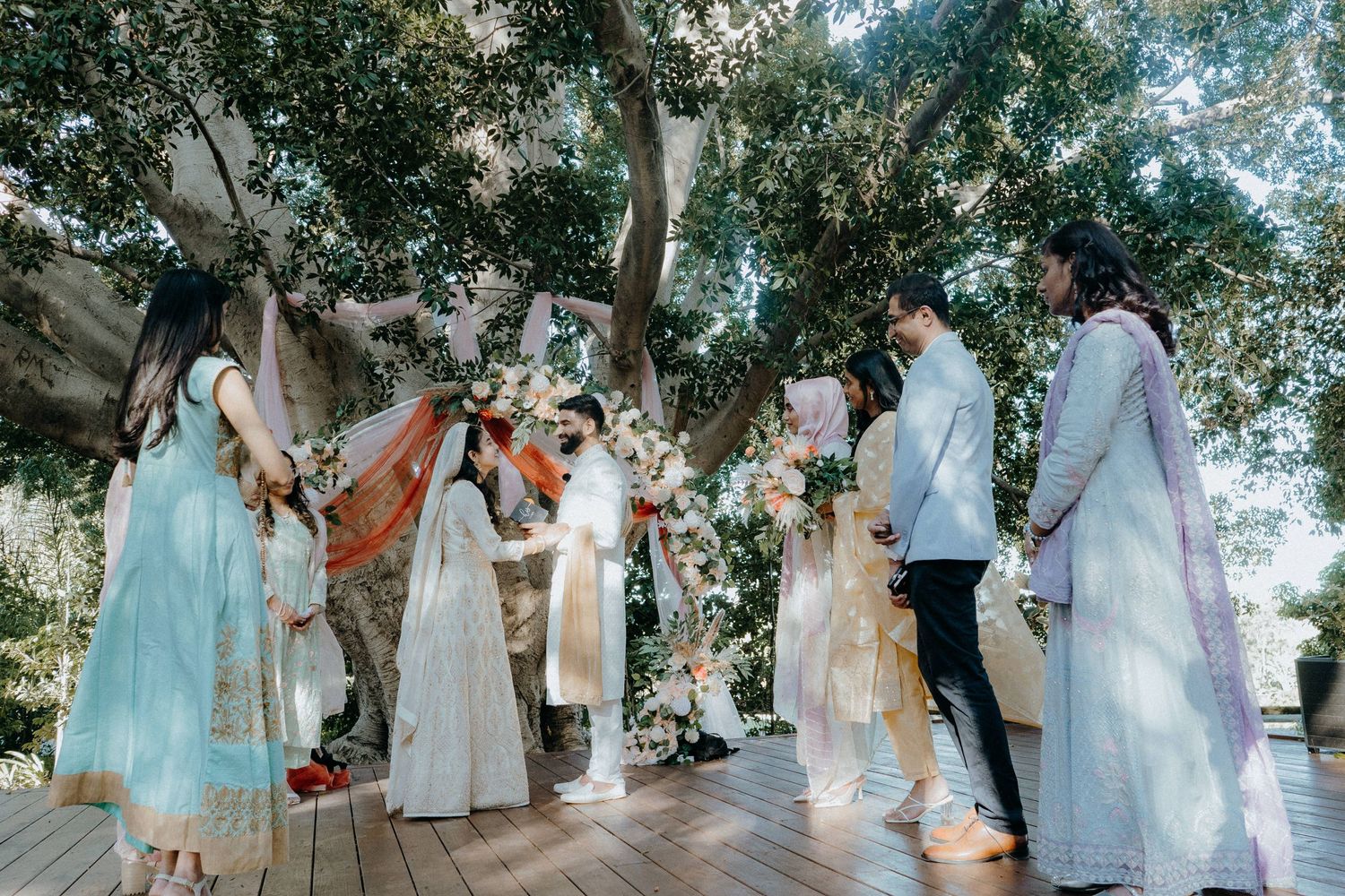 An intimate outdoor wedding ceremony with guests gathered under tree canopy and floral decorations.