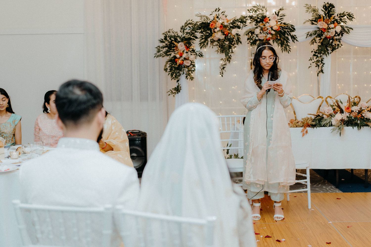 Wedding guest giving a speech at reception with decorative floral arrangements in the background.