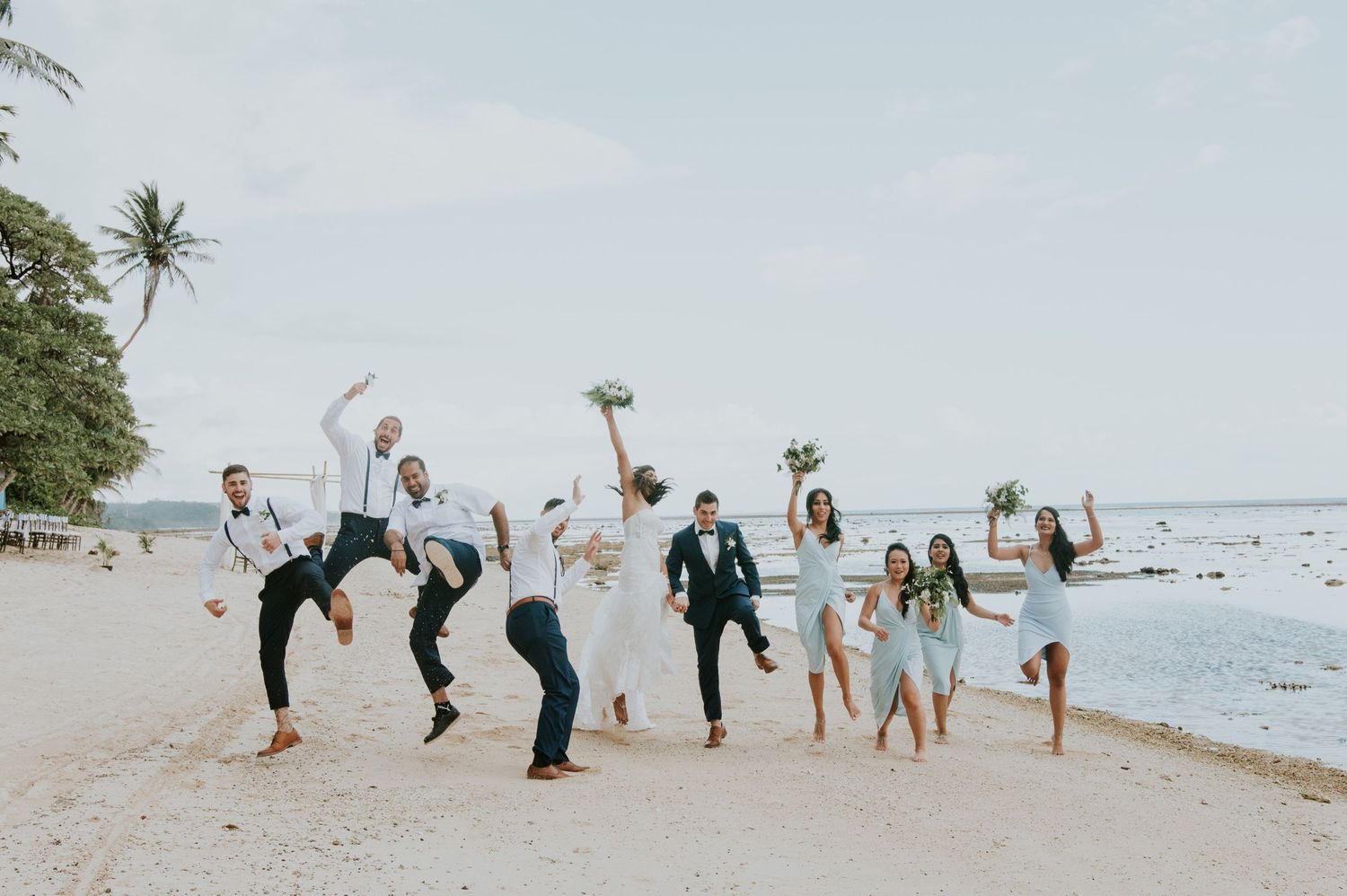 Wedding party jumping and celebrating together on a scenic beach with palm trees.
