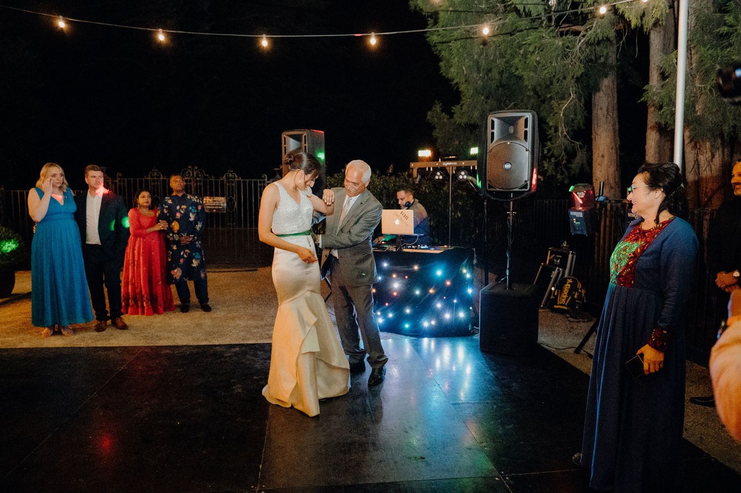 Newlyweds share their first dance near a lit DJ booth at an outdoor evening reception.