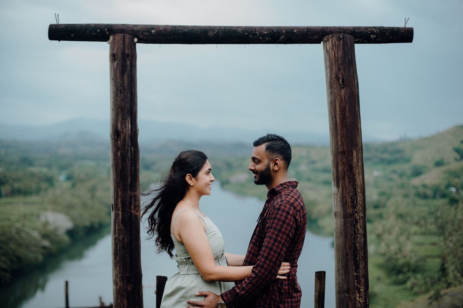 Two people share an intimate moment under a rustic wooden frame with a river vista behind them.