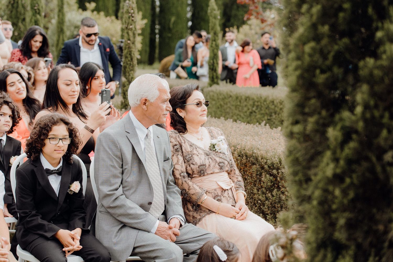 Wedding guests seated in rows at an outdoor ceremony with cypress trees in background.