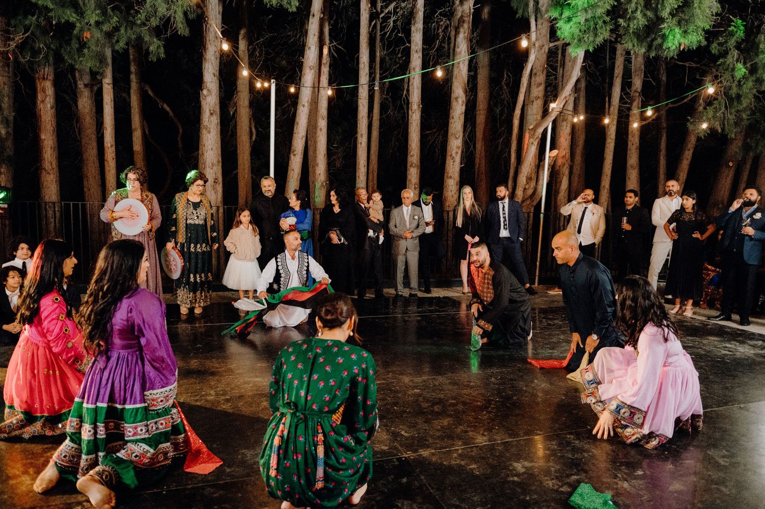Couples dance together on an outdoor dance floor surrounded by tall trees and string lights during a festive celebration.