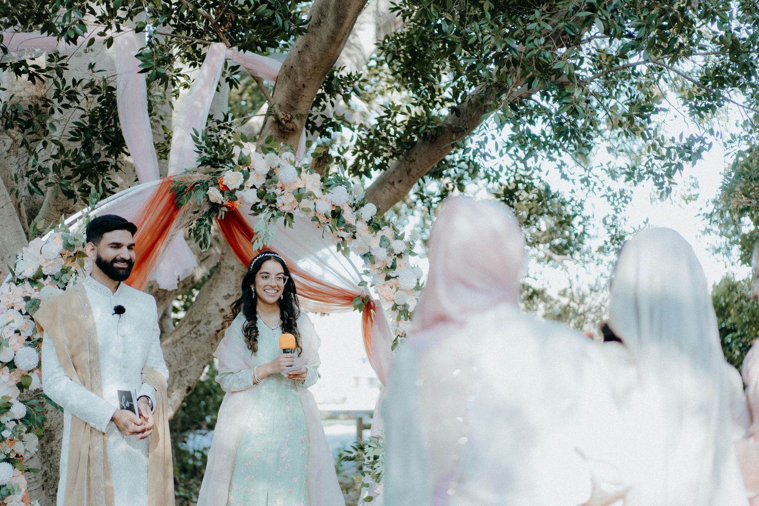 An outdoor wedding ceremony under a tree with soft ethereal lighting and white decorations.