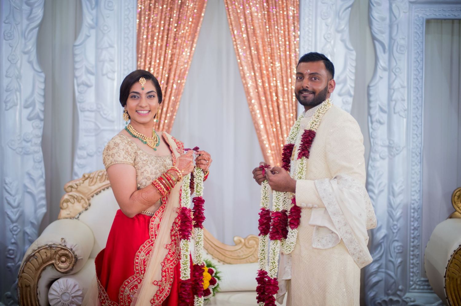 Indian wedding ceremony with couple exchanging garlands in front of decorated white backdrop.