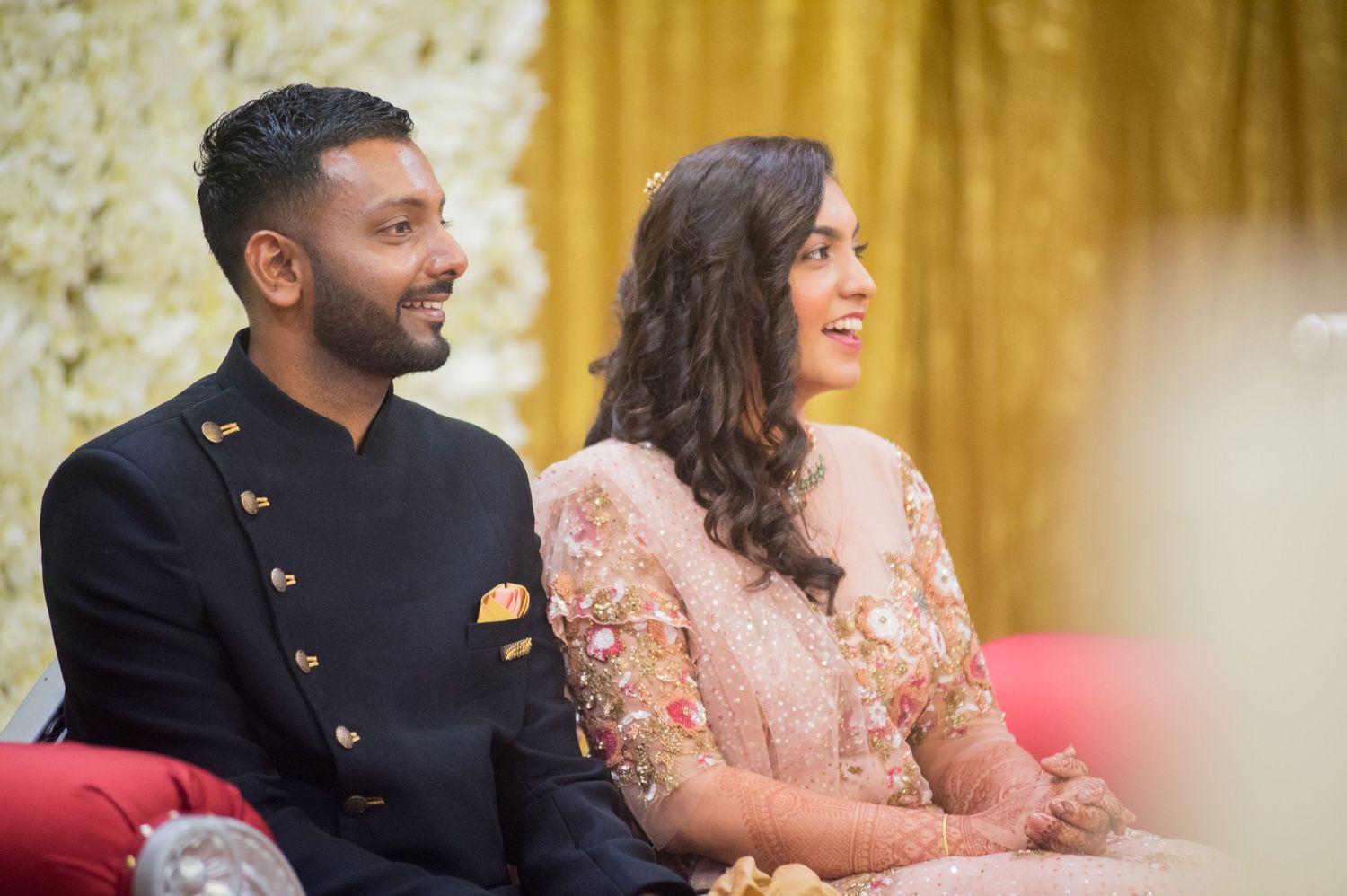 Couple in traditional Indian attire smiling at a celebration event with gold backdrop.