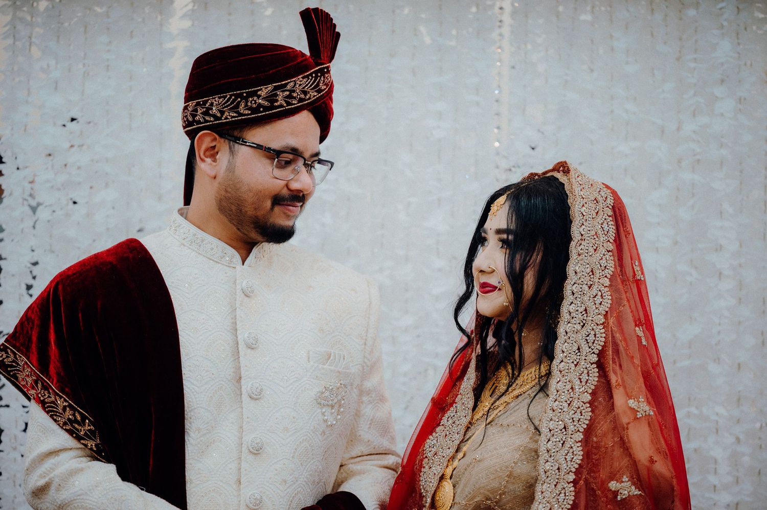 A couple in traditional South Asian wedding attire share a joyful moment against a light background.