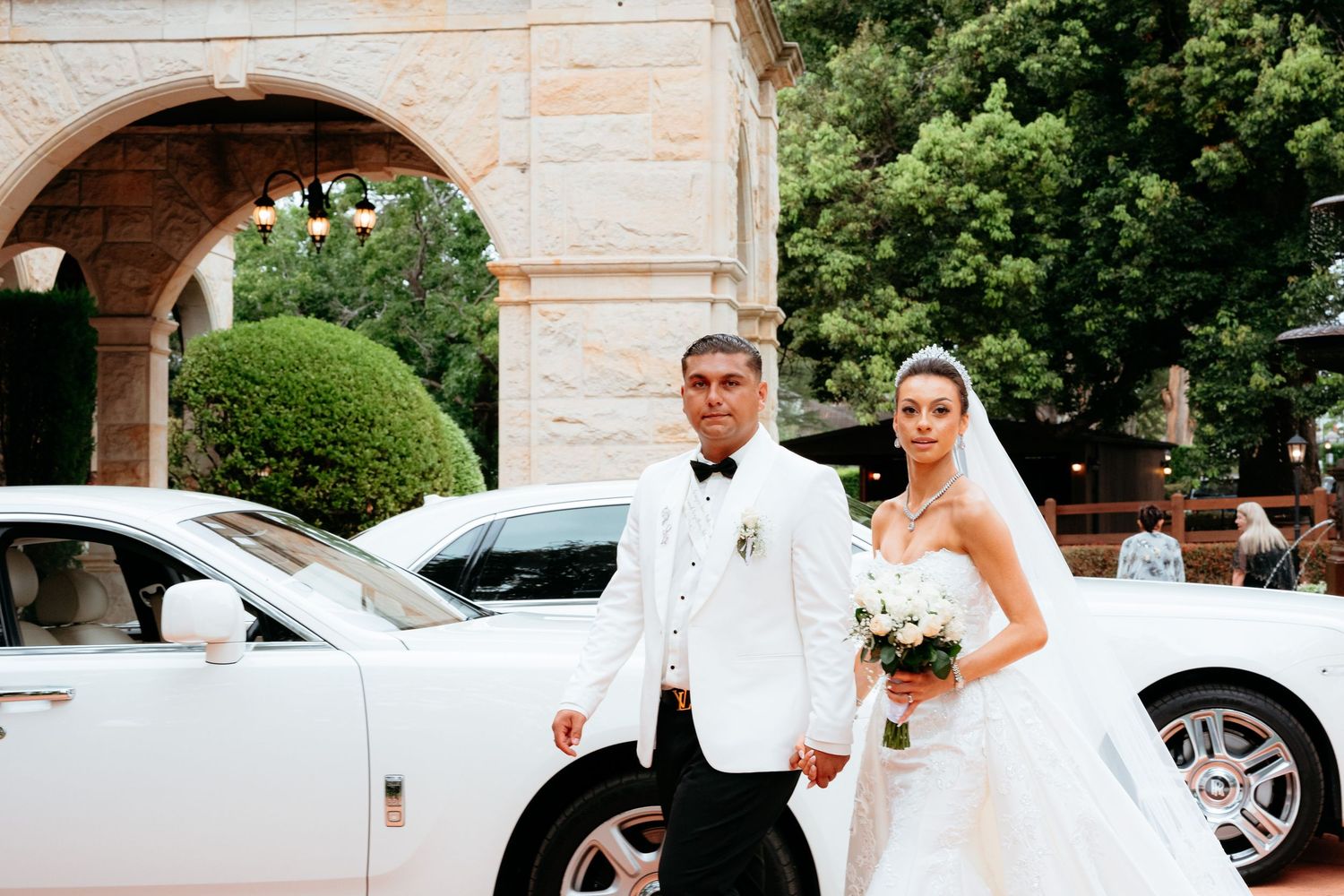 Elegant bride and groom posing beside a white luxury car at a wedding venue with stone archway.