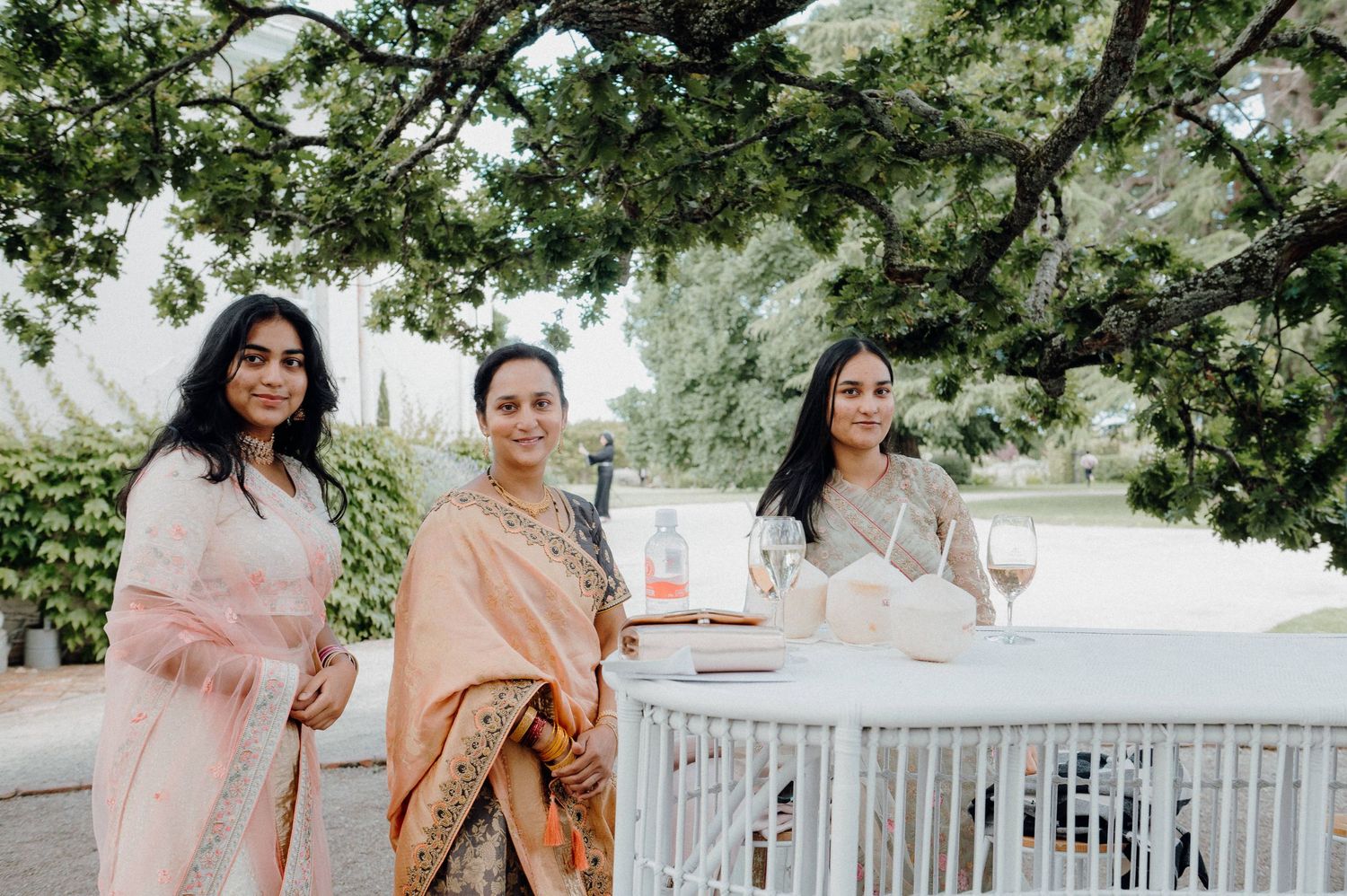 Three guests in soft pink and floral attire pose near white decorative birdcage outdoors.