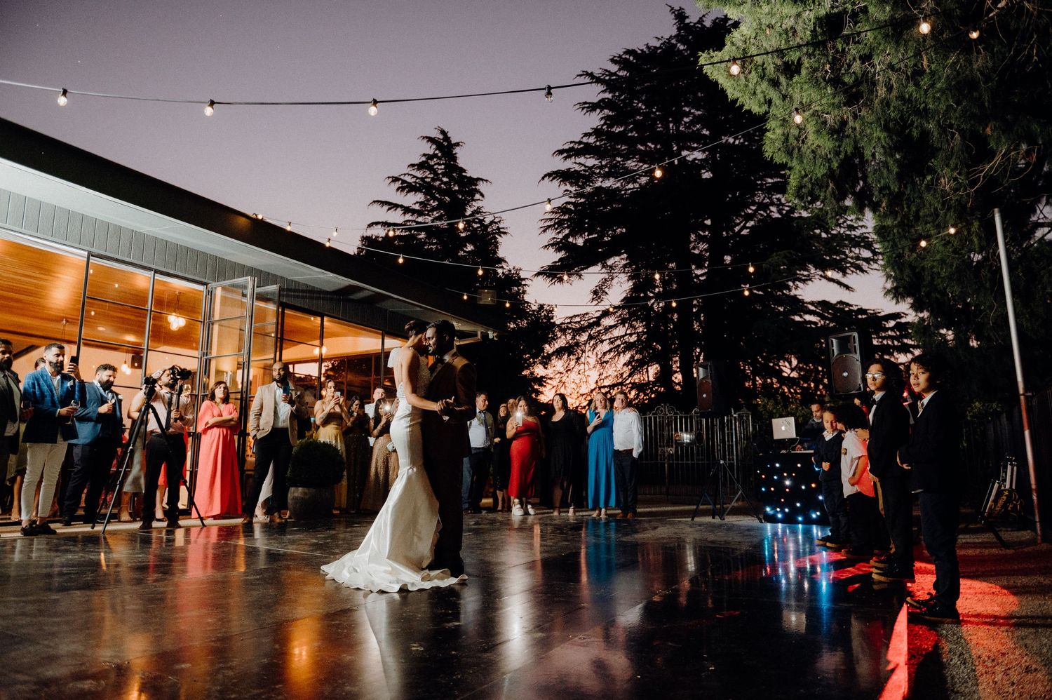 Wedding reception dance floor at twilight with string lights and gathering guests.