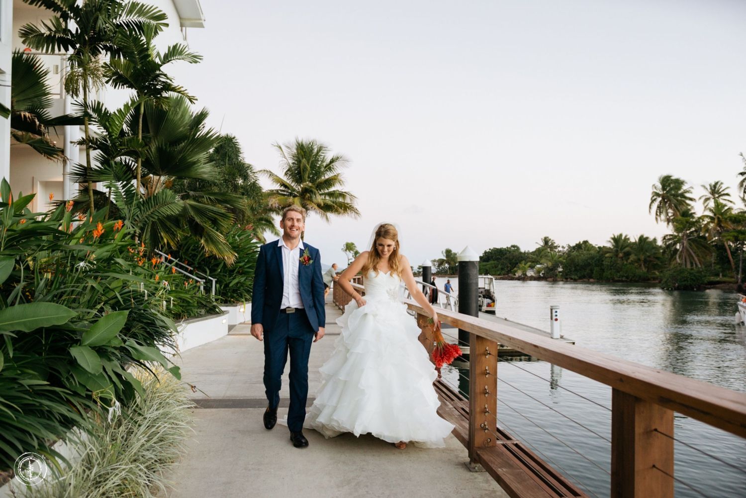 Newlyweds walk along a waterfront path lined with palm trees and boats.
