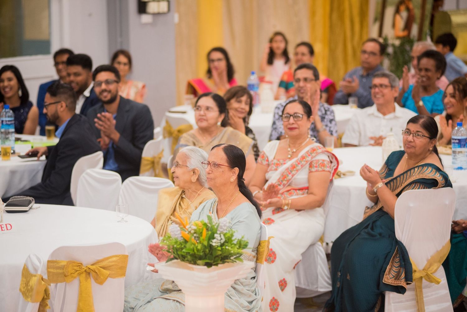 Wedding guests seated in white chairs with gold sashes watching entertainment.
